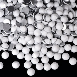 Op de afbeelding: Een stapel witte ronde strass steentjes met een zilveren achterkant. De strass steentjes zijn verspreid over een zwart oppervlak.