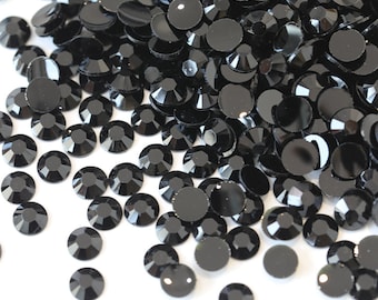 May include: A large quantity of black rhinestones, scattered on a white surface. The rhinestones are small and round, with a faceted design.