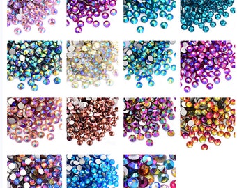 Könnte beinhalten: Sortiment aus schillernden Strasssteinen in verschiedenen Farben, darunter Lila, Blau, Gold und Braun. Die Strasssteine sind in Reihen auf einem weißen Hintergrund angeordnet.