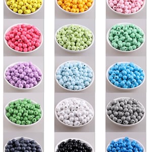 May include: Twelve white bowls filled with colourful wooden beads. The beads are in shades of yellow, orange, pink, red, green, purple, blue, white, grey, navy, black, and turquoise. The round beads have a small hole, ideal for crafting projects.
