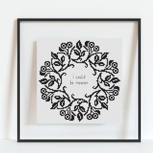 May include: Cross-stitch art framed in black. The artwork features a black floral wreath design with the text "i could be meaner" in the center. The piece is displayed on a white background.