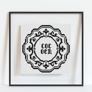 May include: Framed black and white cross-stitch art. The artwork features a decorative, ornate border surrounding the text "COC OEN" in a stylized font. The piece is displayed in a black frame against a white background.