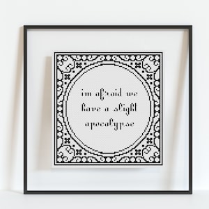 May include: Black and white cross-stitch embroidery framed in a black frame. The embroidery features a decorative border and the text "I'm afraid we have a slight apocalypse."