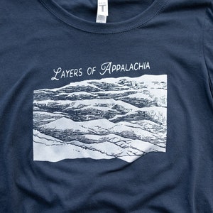 以下が含まれることがあります： 紺色のTシャツに、白い山脈のグラフィックと「Layers of Appalachia」というテキストがプリントされています。