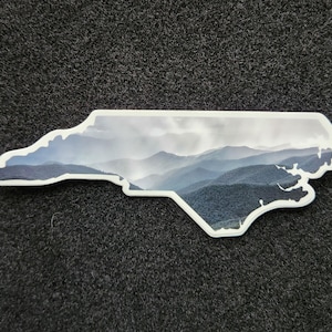 May include: A sticker in the shape of North Carolina, featuring a scenic mountain landscape in shades of blue and gray. The sticker has a white border and is set against a dark gray textured background.