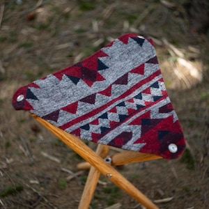 May include: A wooden folding stool with a red, gray, and black patterned fabric seat. The fabric has a geometric design with triangles and diamonds.