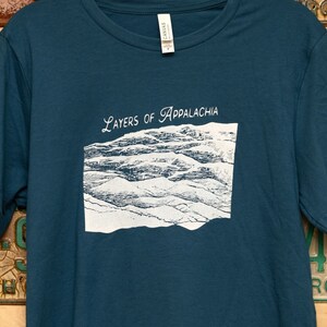 Pode incluir: Uma camiseta azul turquesa com um gráfico branco de montanhas e o texto "Layers of Appalachia".