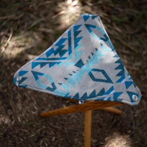 May include: A triangular folding stool with a gray and blue patterned seat. The seat has a geometric design in shades of blue and gray. The wooden legs are angled for stability, and the stool is set outdoors.