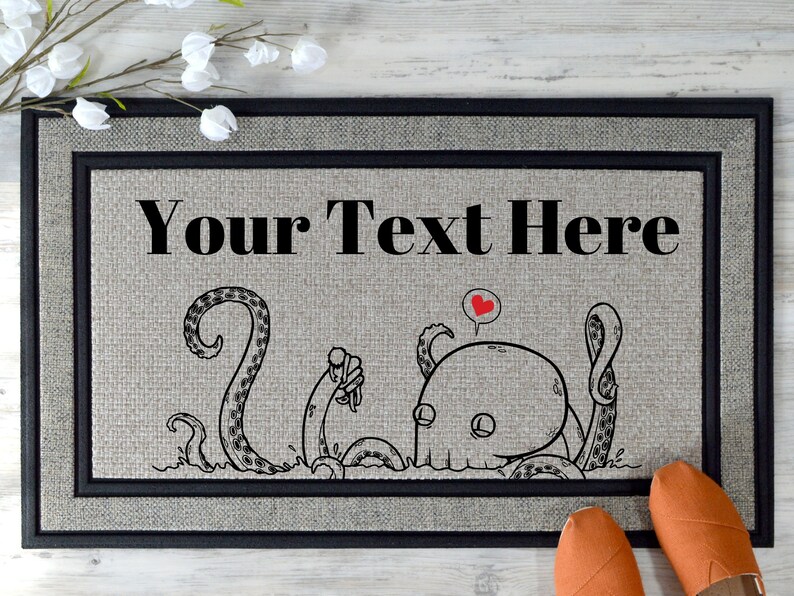 Pu&ograve; includere: Un tappetino per la porta con un bordo nero e uno sfondo grigio. Il tappetino presenta un'illustrazione in bianco e nero di un polpo con un cuore sopra la testa. Il testo "Your Text Here" &egrave; stampato in nero nella parte superiore del tappetino.