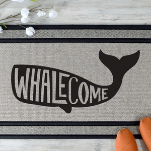 May include: A black and white doormat with a whale silhouette. The word "WHALEcome" is spelled out inside the whale's body.
