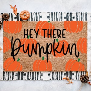 Op de afbeelding: Een bruine deurmat met de tekst "Hey there pumpkin" in zwarte letters. De deurmat is versierd met oranje pompoenen met groene stelen.