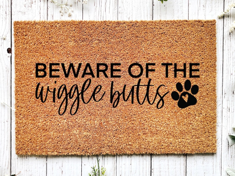 May include: A brown coir doormat with the text "BEWARE OF THE wiggle butts" in black lettering and a black paw print with a heart inside.