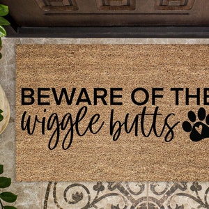 May include: A brown coir doormat with the text "BEWARE OF THE wiggle butts" in black lettering and a black paw print with a heart inside.