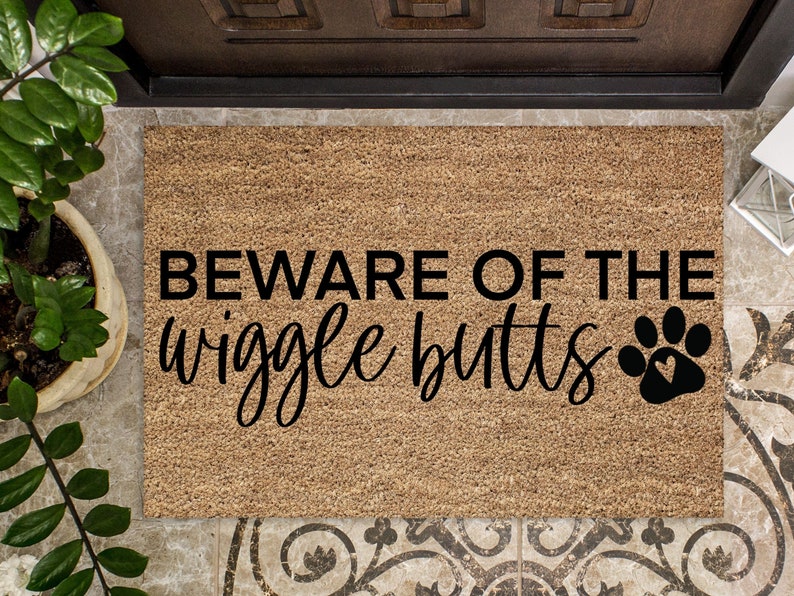 May include: A brown coir doormat with the text "BEWARE OF THE wiggle butts" in black lettering and a black paw print with a heart inside.