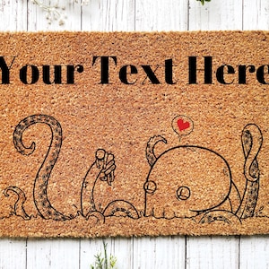 Pu&ograve; includere: Un tappetino per porta in fibra di cocco marrone con il testo "Your Text Here" in lettere nere. Il tappetino presenta un'illustrazione in bianco e nero di due polpi dei cartoni animati, uno con un fumetto a forma di cuore.