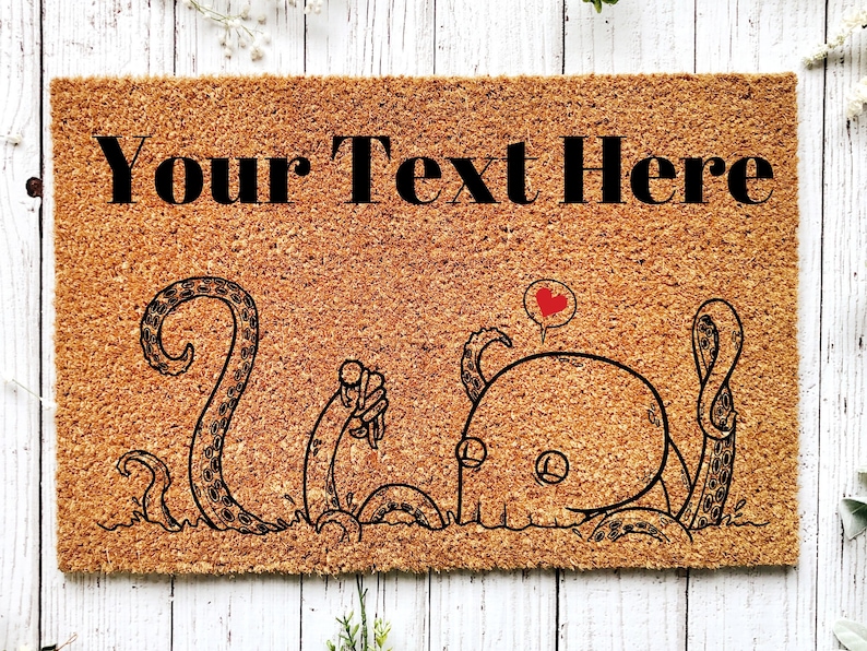 Pu&ograve; includere: Un tappetino per porta in fibra di cocco marrone con il testo "Your Text Here" in lettere nere. Il tappetino presenta un'illustrazione in bianco e nero di due polpi dei cartoni animati, uno con un fumetto a forma di cuore.