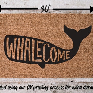 May include: A brown coir doormat with the word "WHALECOME" printed in black lettering inside the silhouette of a whale. The doormat measures 30 inches wide by 18 inches tall.