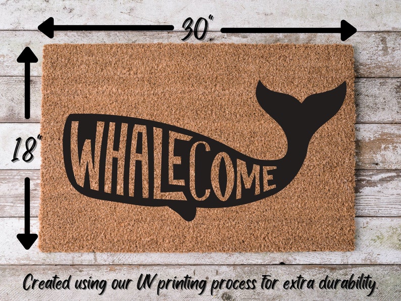 May include: A brown coir doormat with the word "WHALECOME" printed in black lettering inside the silhouette of a whale. The doormat measures 30 inches wide by 18 inches tall.