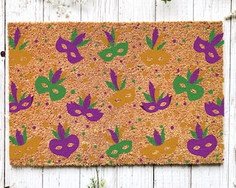May include: A brown coir doormat with a repeating pattern of purple, green, and gold Mardi Gras masks. The masks have feathers and are set against a background of small gold and green dots.