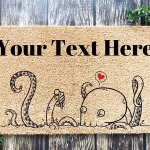 Pu&ograve; includere: Un tappetino per porta in fibra di cocco marrone con il testo "Your Text Here" in lettere nere. Un'illustrazione di cartone animato di due polpi, uno con un cuore sopra la testa, &egrave; stampato sul tappetino.