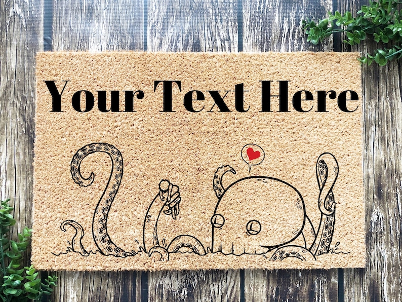 Pu&ograve; includere: Un tappetino per porta in fibra di cocco marrone con il testo "Your Text Here" in lettere nere. Un'illustrazione di cartone animato di due polpi, uno con un cuore sopra la testa, &egrave; stampato sul tappetino.