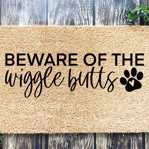 May include: A brown coir doormat with the text "BEWARE OF THE wiggle butts" in black lettering and a black paw print with a heart inside.