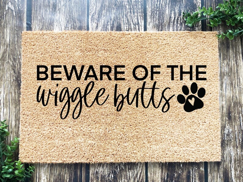 May include: A brown coir doormat with the text "BEWARE OF THE wiggle butts" in black lettering and a black paw print with a heart inside.