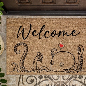Pu&ograve; includere: Un tappetino per porta in fibra di cocco marrone con la parola "Welcome" in lettere corsive nere. Sotto il testo c'&egrave; un'illustrazione in bianco e nero di un polpo con un cuore rosso sopra la testa.