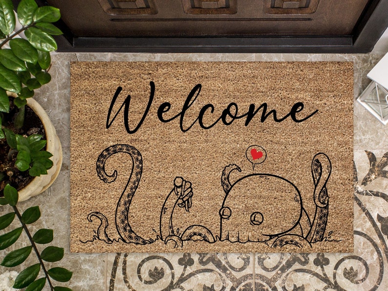 Pu&ograve; includere: Un tappetino per porta in fibra di cocco marrone con la parola "Welcome" in lettere corsive nere. Sotto il testo c'&egrave; un'illustrazione in bianco e nero di un polpo con un cuore rosso sopra la testa.