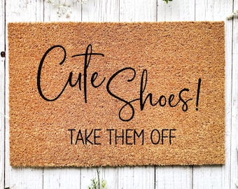 May include: A rectangular doormat made of natural coir fiber. The mat features the words "Cute Shoes! TAKE THEM OFF" in a black, elegant script. The mat is placed on a white wooden surface.