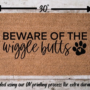 May include: A brown coir doormat with the text "BEWARE OF THE wiggle butts" and a black paw print with a heart in the center. The doormat measures 30 inches wide by 18 inches tall.