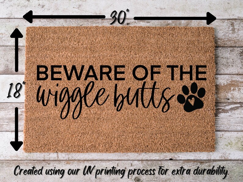 May include: A brown coir doormat with the text "BEWARE OF THE wiggle butts" and a black paw print with a heart in the center. The doormat measures 30 inches wide by 18 inches tall.