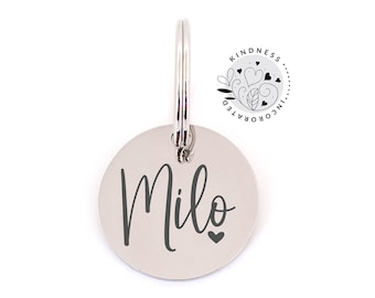 May include: A silver-colored, circular pet tag with the name "Milo" in a dark gray script font and a small heart. The tag has a silver ring for attachment and a small logo that says "Kindness Incorporated".