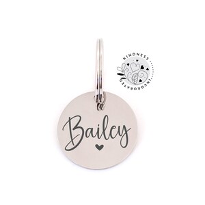 May include: A silver-colored, circular pet tag with the name "Bailey" in a cursive font and a small heart symbol. The tag is attached to a metal ring. The upper right corner features a circular logo with the text "Kindness Incorporated".