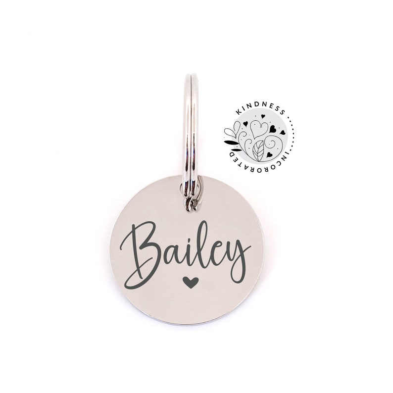 May include: A silver-colored, circular pet tag with the name "Bailey" in a cursive font and a small heart symbol. The tag is attached to a metal ring. The upper right corner features a circular logo with the text "Kindness Incorporated".