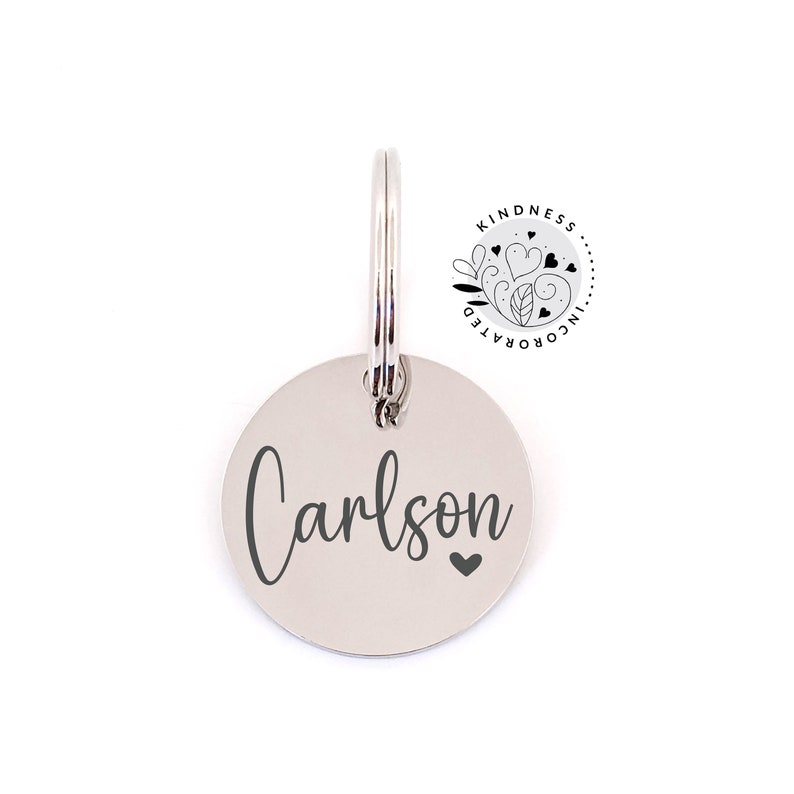 May include: A silver-colored, circular pet tag with the name "Carlson" engraved in a cursive font, accompanied by a small heart symbol. The tag is attached to a silver ring. The upper right corner features a logo with the text "Kindness Incorporated".