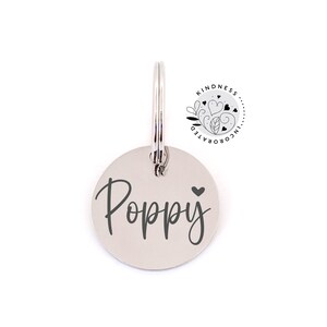 May include: A round, silver pet tag with the name "Poppy" in a cursive font and a small heart symbol. The tag is attached to a silver ring. The upper right corner features a circular logo with the words "Kindness Incorporated".