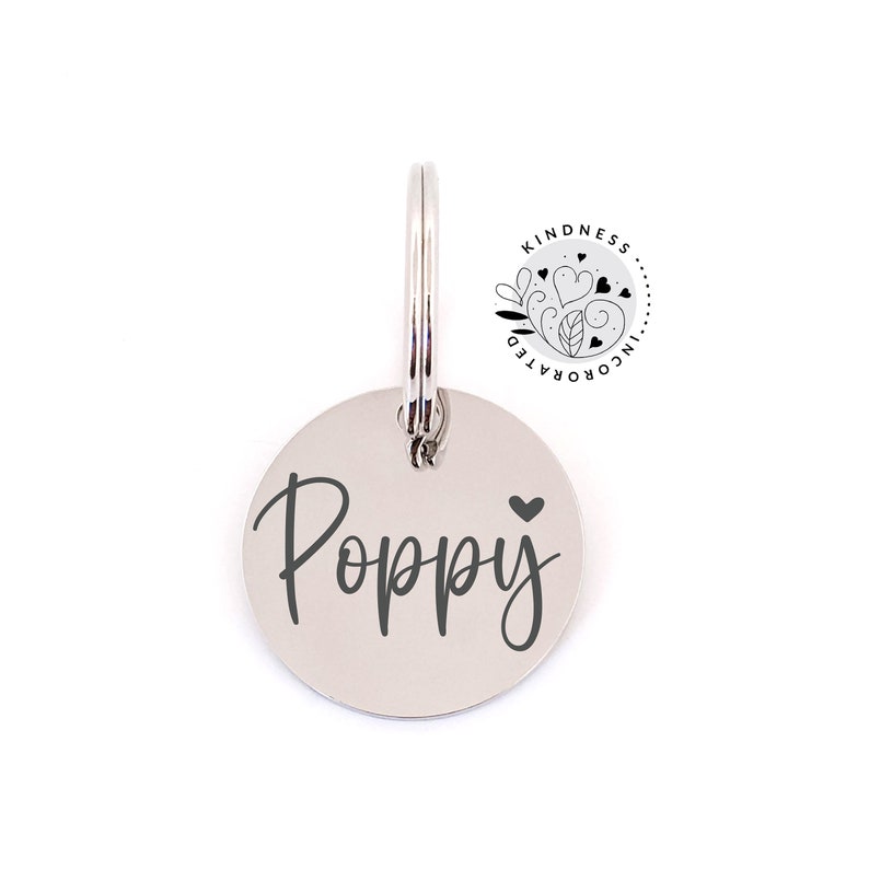 May include: A round, silver pet tag with the name "Poppy" in a cursive font and a small heart symbol. The tag is attached to a silver ring. The upper right corner features a circular logo with the words "Kindness Incorporated".