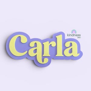 Puede incluir: Un recorte de nombre en capas en amarillo y morado. El nombre "Carla" está en una fuente en negrita y redondeada, con un color amarillo. El nombre está superpuesto sobre un fondo morado. El logotipo de Kindness Inc. está en la esquina superior derecha.