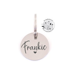 May include: A silver-colored, circular pet tag with the name "Frankie" in a script font and a small heart below the name. The tag is attached to a metal ring. A circular logo with the text "Kindness Incorporated" is in the upper right corner.