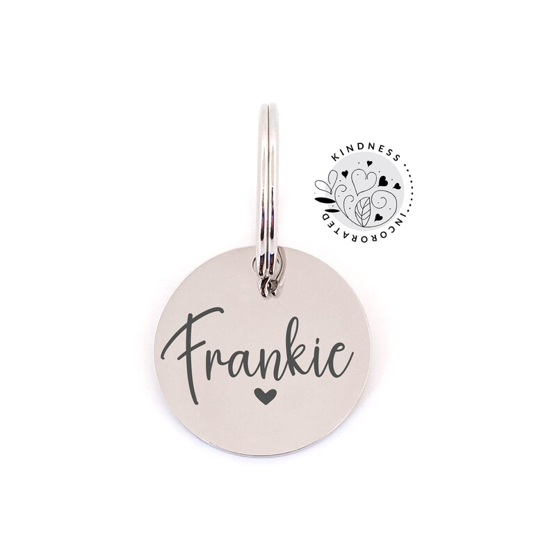 May include: A silver-colored, circular pet tag with the name "Frankie" in a script font and a small heart below the name. The tag is attached to a metal ring. A circular logo with the text "Kindness Incorporated" is in the upper right corner.