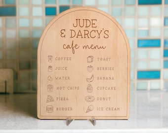 May include: A wooden sign with a rounded top, featuring the text "JUDE & DARCY'S cafe menu" in a handwritten font. The sign lists various food and drink items with corresponding icons, including coffee, toast, juice, berries, water, banana, hot chips, cupcake, pizza, donut, burger, and ice cream.