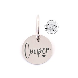 May include: A silver-colored, circular pet tag with the name "Cooper" in a cursive font and a small heart symbol. The tag is attached to a metal ring. The Kindness Incorporated logo is visible in the upper right corner.