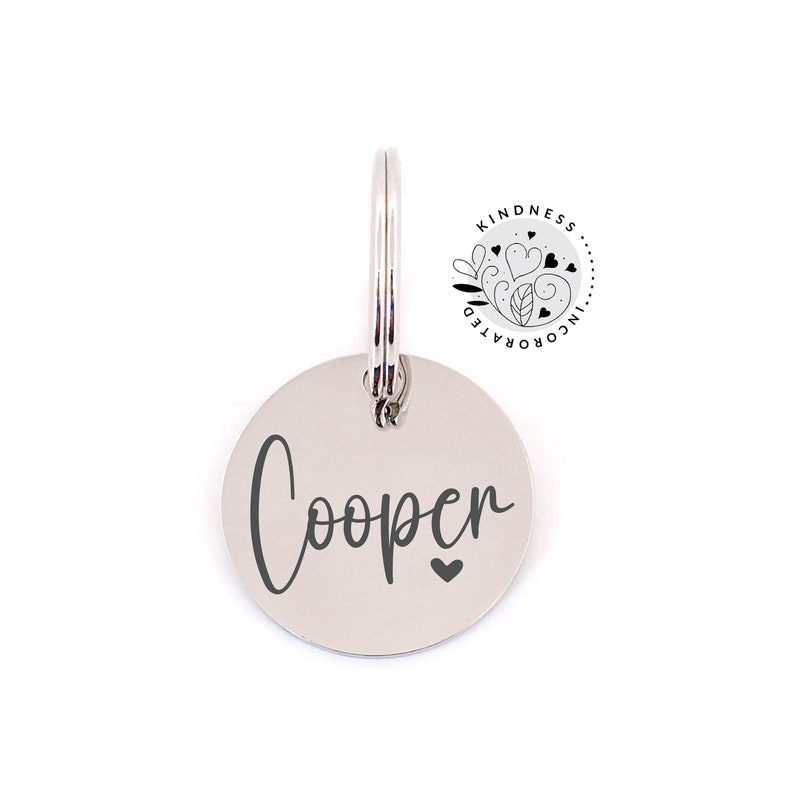 May include: A silver-colored, circular pet tag with the name "Cooper" in a cursive font and a small heart symbol. The tag is attached to a metal ring. The Kindness Incorporated logo is visible in the upper right corner.