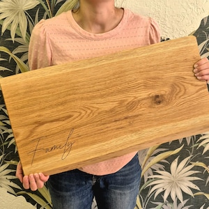 May include: A rectangular wooden cutting board with the word "Family" in a cursive font. The board is made of light-colored wood with visible grain patterns. The board is held by a person.