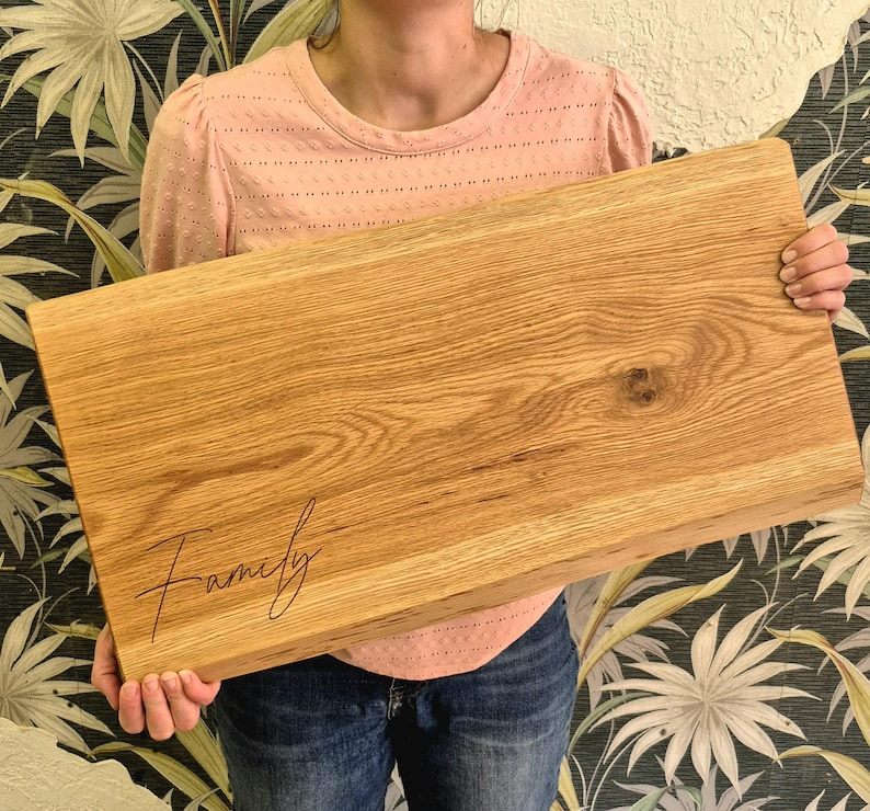 May include: A rectangular wooden cutting board with the word "Family" in a cursive font. The board is made of light-colored wood with visible grain patterns. The board is held by a person.