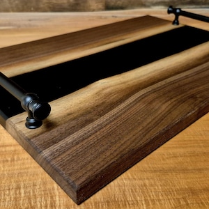 May include: A rectangular wooden serving tray with a black central stripe and black metal handles. The tray is made of dark brown wood with a natural grain pattern. The handles are attached to the sides.