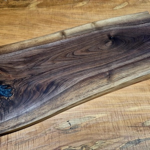 May include: A rectangular wooden serving board with a live edge. The board is made of dark brown walnut wood with a natural wood grain pattern. A section of blue resin fills a knot in the wood. The board rests on a light brown wooden surface.
