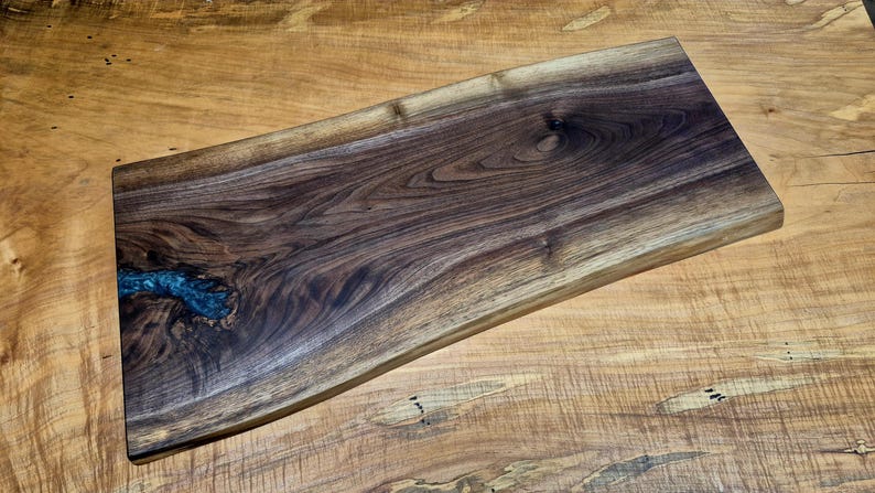 May include: A rectangular wooden serving board with a live edge. The board is made of dark brown walnut wood with a natural wood grain pattern. A section of blue resin fills a knot in the wood. The board rests on a light brown wooden surface.