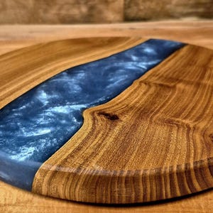 May include: A round wooden serving board with a dark blue epoxy resin river design. The wood has a warm, natural tone, and the resin has a glossy finish. The board is ideal for serving cheese, charcuterie, or other appetizers.